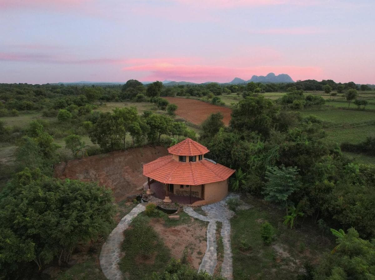 BhuDeep from above at dusk — a round, tiled-roof earthen villa among scrub, fields and distant hills